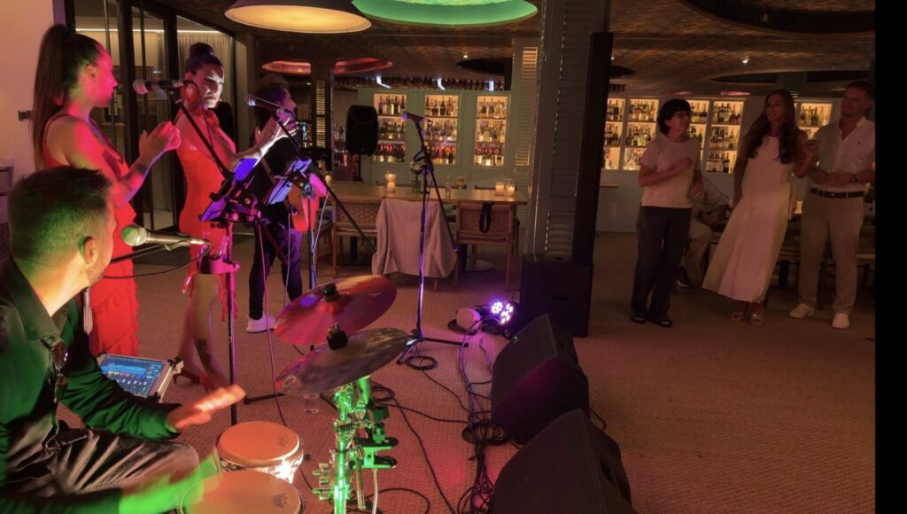 In einer warm beleuchteten Bar spielt eine kleine Band: links ein Percussionist an Handtrommeln, daneben zwei Saengerinnen mit Mikrofonen und klatschenden Haenden; rechts tanzen und schauen Gaeste zu. Die Szene passt zu Rumba Fusion und Pop Flamenco fuer Firmenevents in Barcelona.