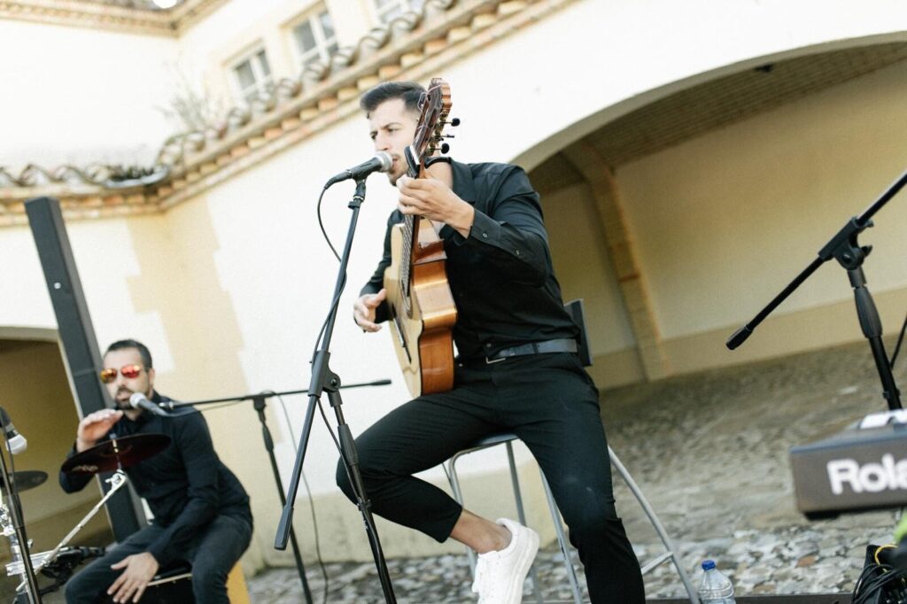 Músico sentado num banco a cantar num microfone enquanto toca guitarra espanhola, com um percussionista ao lado e equipamento de som, num pátio exterior com arcos; ambiente descontraído de flamenquito e pop flamenco em Barcelona para aniversário com bom astral.