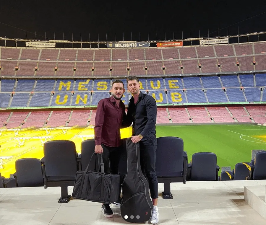 Dos hombres posan en la grada de un gran estadio de fútbol vacío, con el césped al fondo. Delante de ellos hay varias butacas azules y cada uno sostiene una funda de instrumento: una de guitarra acústica rígida y otra bolsa grande de equipo. Imagen asociada a flamenquito y pop flamenco en Barcelona para cumpleaños con buen rollo, estilo Rumba con Alma.
