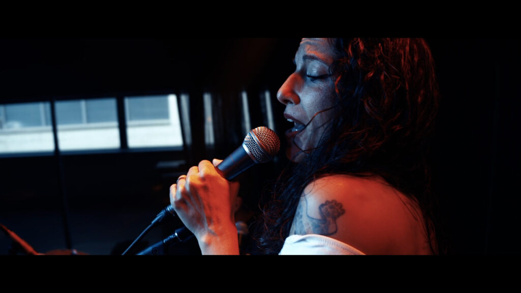 Cantora de perfil a segurar um microfone com as duas mãos, a cantar com os olhos fechados num ambiente interior escuro e luz quente, sugerindo rumba fusão e pop flamenco para eventos de empresa em Barcelona