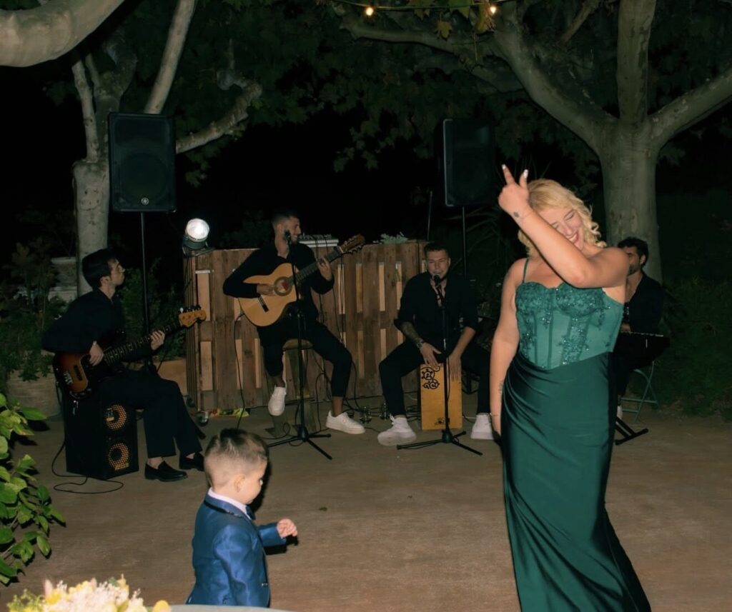 En un jardí de nit, una dona amb vestit verd balla somrient davant d’un grup de músics amb guitarra, baix i caixó, amb altaveus als laterals; l’escena transmet bon rotllo i encaixa amb flamenquito i pop flamenc a Barcelona per a aniversaris i celebracions.