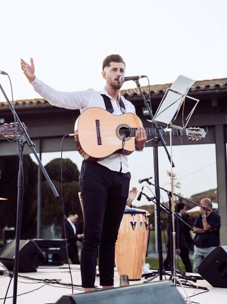 Cantant amb camisa blanca i guitarra espanyola davant d’un micròfon, amb el braç aixecat com si presentés una cançó; a l’escenari hi ha peus de micro i un faristol, amb un calaix de percussió al fons i algunes persones mirant en un espai exterior, ideal per a flamenquito i pop flamenc a Barcelona per a aniversaris amb bon rotllo.