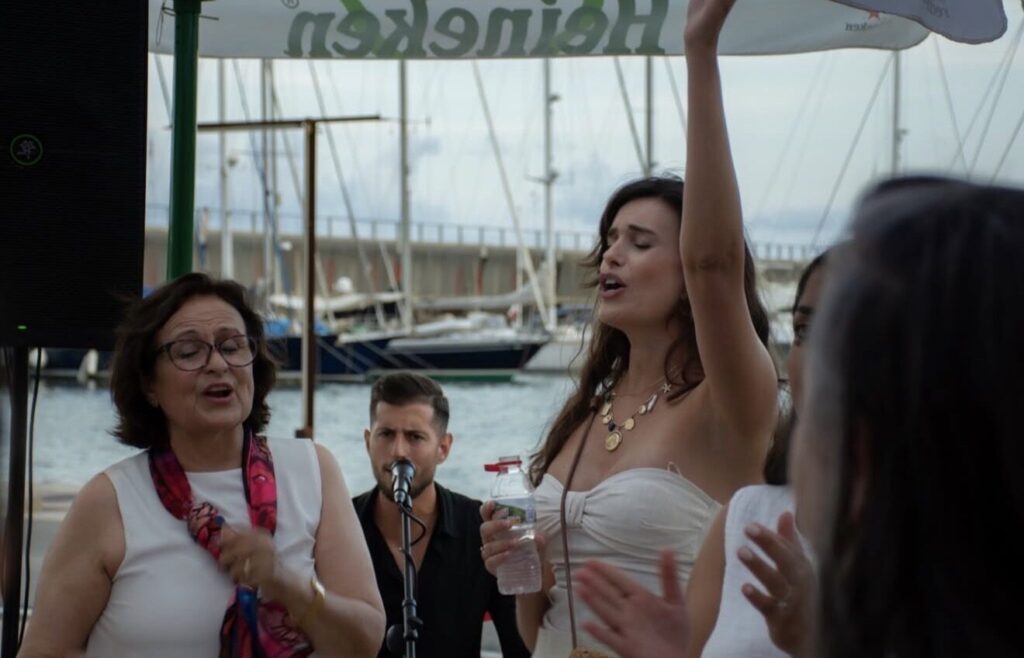 En una terrassa a tocar del port, dues dones canten i aplaudeixen amb el públic mentre un músic amb micròfon queda al fons; l’escena transmet bon rotllo i festa, ideal per a flamenquito i pop flamenc a Barcelona en un aniversari o celebració privada.