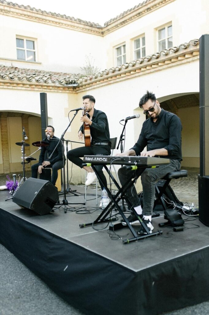 Tres músics en un petit escenari a l’aire lliure, davant d’un edifici de parets clares i teulada de teules. Un toca el teclat amb ulleres de sol, un altre canta i toca una guitarra assegut a un tamboret, i el tercer fa percussió amb un cajón. A l’escena hi ha micròfons, altaveus i cables, en un ambient proper ideal per a flamenquito i pop flamenc a Barcelona per a un aniversari amb bon rotllo.