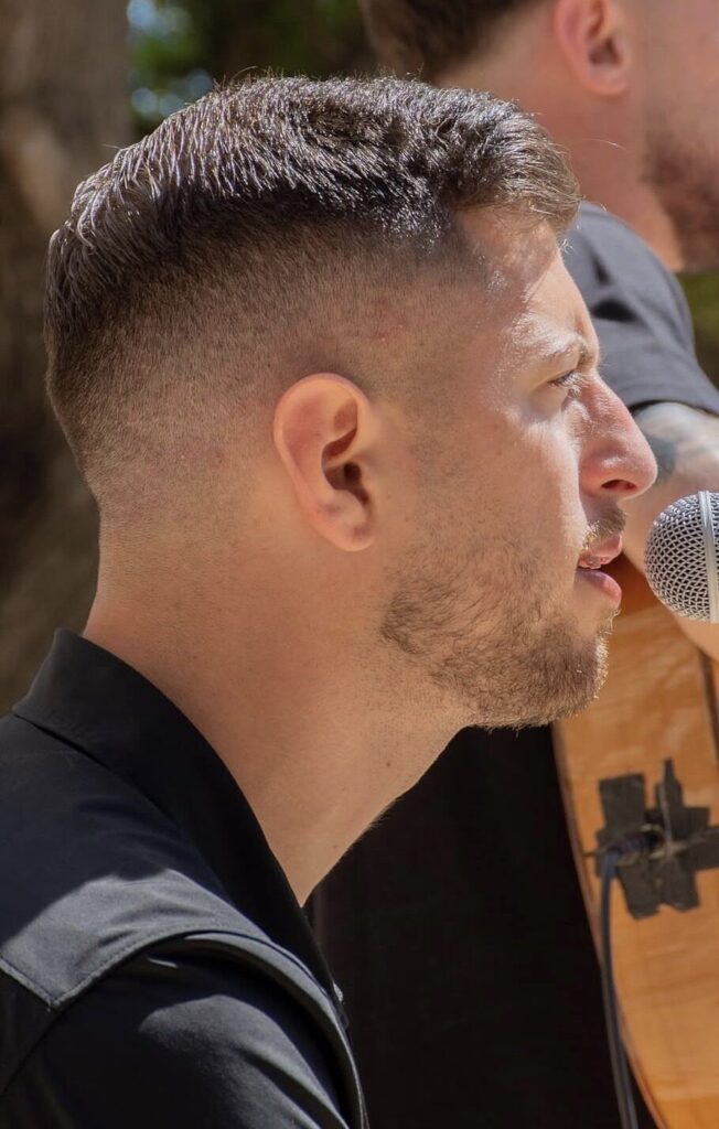 Primer pla de perfil d’un cantant amb camisa negra mentre canta a tocar d’un micròfon; al fons es veu parcialment una guitarra. La escena transmet bon rotllo, ideal per a flamenquito i pop flamenc a Barcelona per a aniversaris.