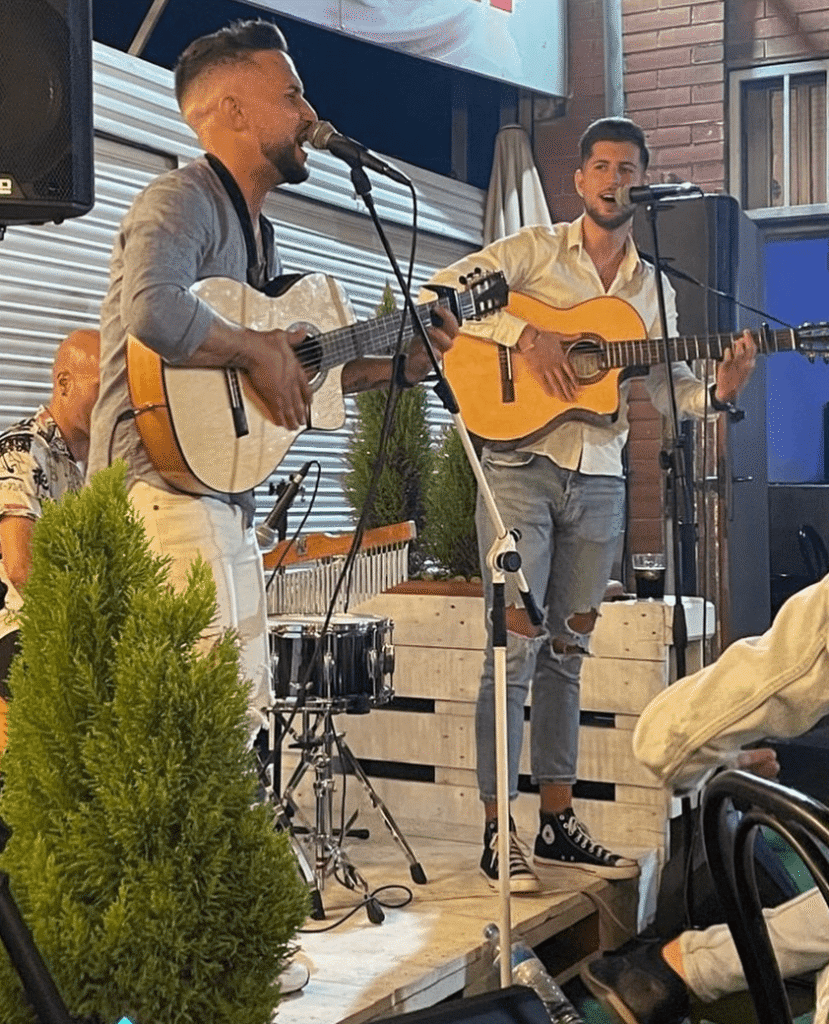 Dos músics toquen guitarres espanyoles i canten a dos micròfons sobre un escenari petit, amb una bateria al darrere i plantes al davant. L’escena transmet bon rotllo i energia, ideal per a flamenquito i pop flamenc a Barcelona per a aniversaris i celebracions.