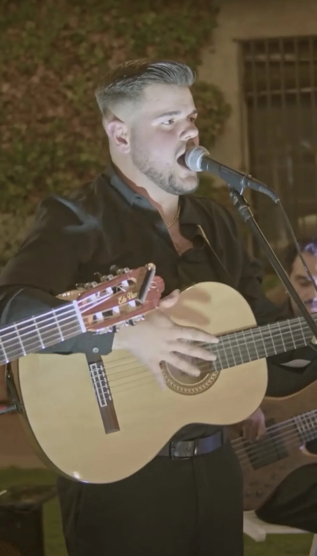 Primer plano de un cantante con camisa negra cantando a un micrófono mientras toca una guitarra española; al fondo se ven plantas y parte de otro músico, en un ambiente de rumba y flamenquito para una boda con fiesta en Barcelona.