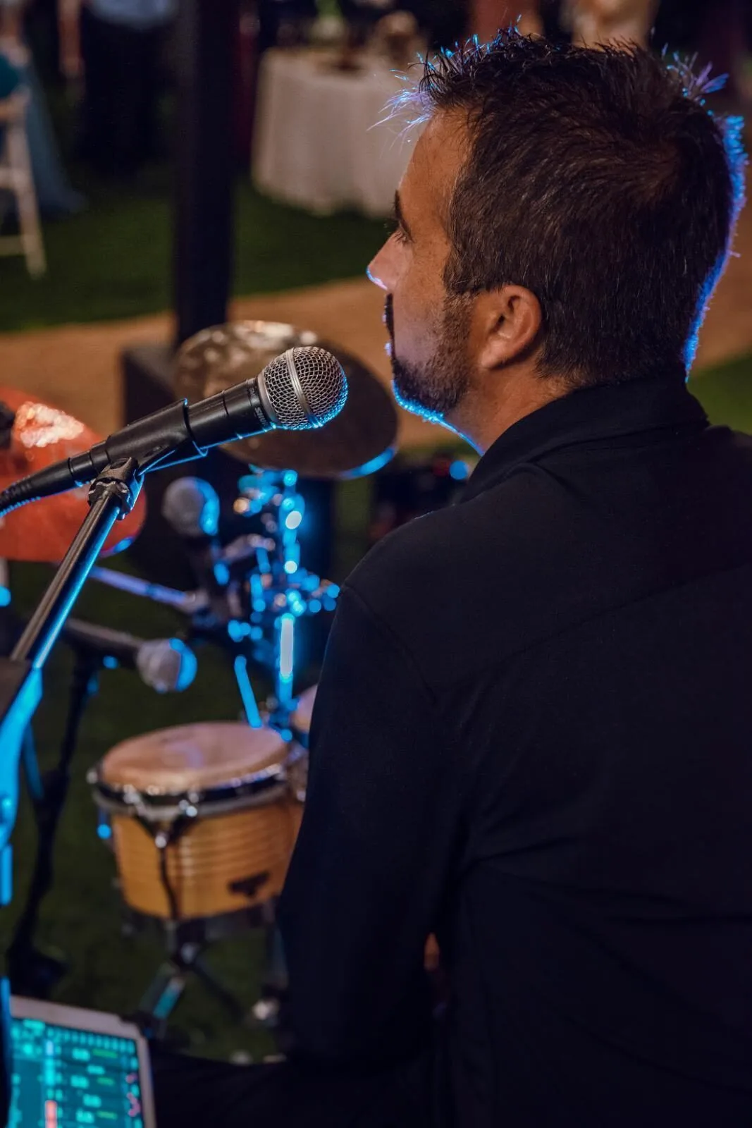 Primer plano de un músico de perfil, vestido de negro, cantando o hablando frente a un micrófono en una celebración; al fondo se ven una conga y parte de la batería desenfocadas. La escena sugiere rumba y flamenquito para una boda con fiesta en Barcelona, con iluminación cálida y ambiente de evento privado.