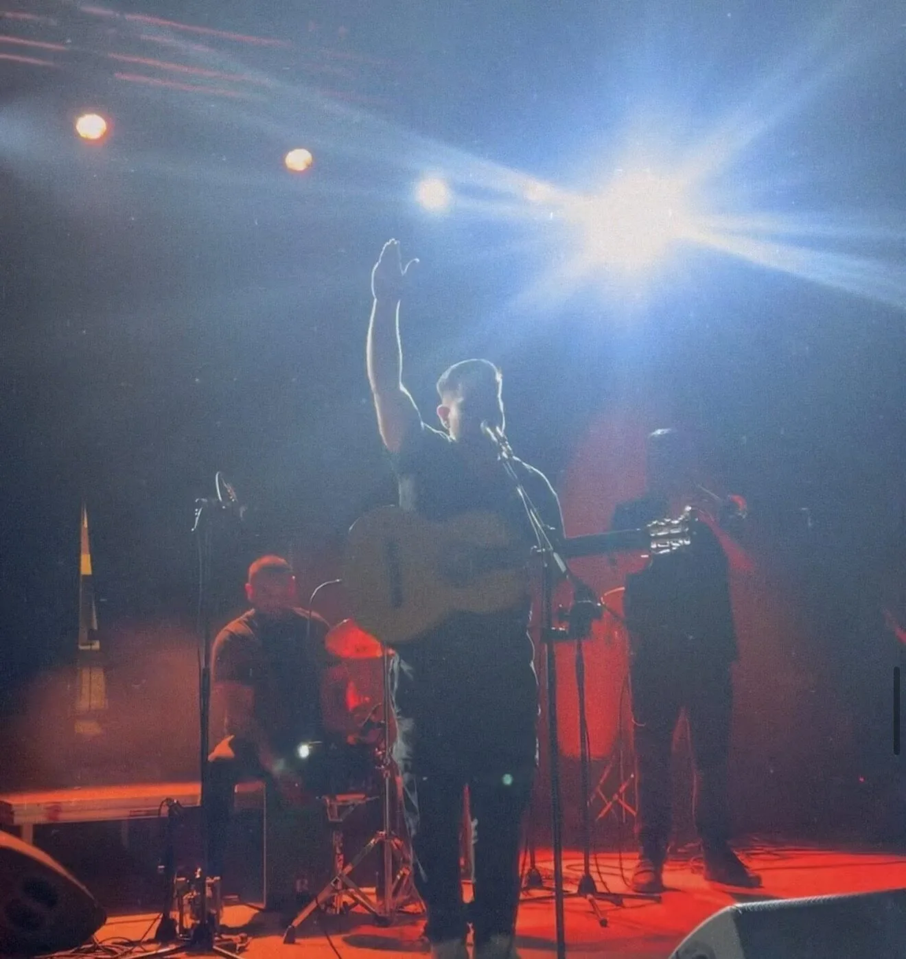 En un escenario con luces rojas y un foco blanco muy intenso, un músico canta al micrófono mientras toca una guitarra española y levanta una mano; detrás se distingue un percusionista sentado y, a la derecha, otro guitarrista en sombra, creando un ambiente de rumba y flamenquito para una boda con fiesta en Barcelona.