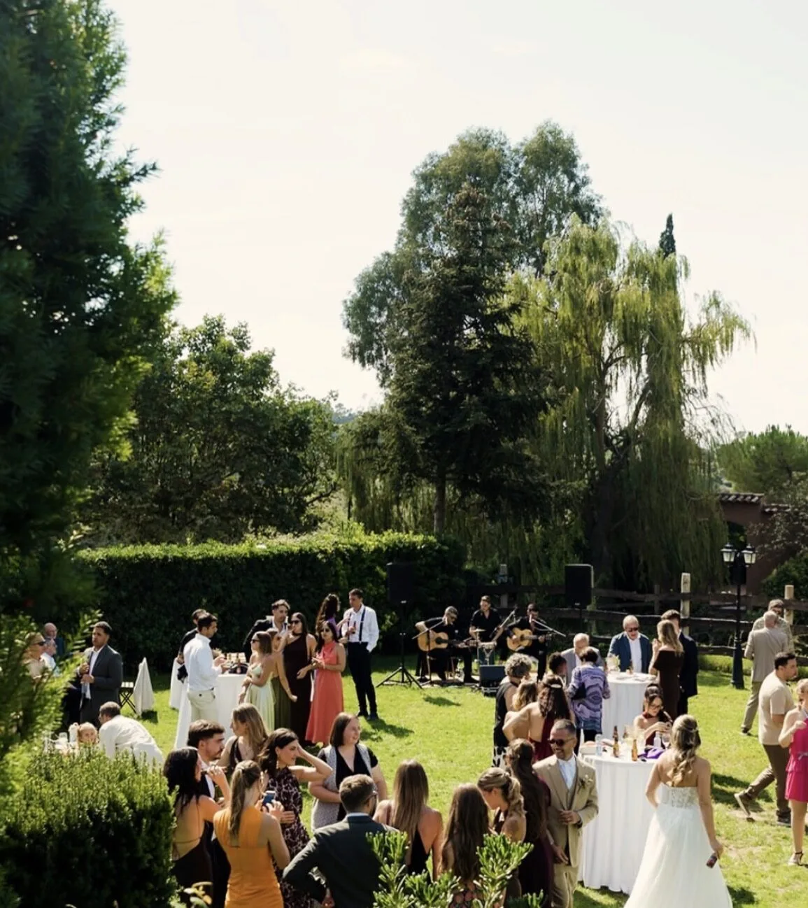 Invitados reunidos en un jardín durante un cóctel de boda, con varias mesas altas y un grupo de músicos al fondo tocando con guitarras y altavoces; ambiente de rumba y flamenquito para una boda con fiesta en Barcelona.