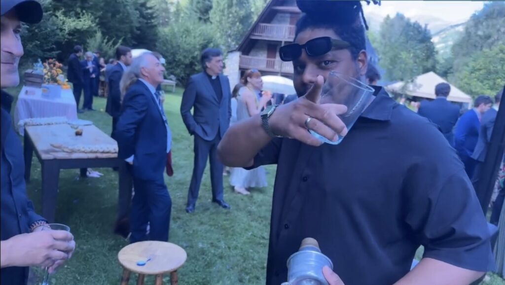 户外花园酒会场景中，一名穿深色衬衫的男子靠近镜头，举着透明杯子并用手指向前方，头顶夹着太阳镜；背景里有穿西装的宾客在草地上交谈，旁边摆着点心与饮品的长桌，远处可见木屋与树木，呈现适合在巴塞罗那夜晚营造加泰罗尼亚风格西班牙欢快音乐与都市气息的派对氛围