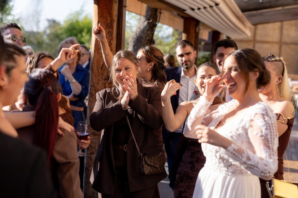 户外木质露台的婚礼宾客围在一起跳舞和鼓掌，前景一位穿白色蕾丝礼服的新娘笑着举起手，旁边的宾客也跟着挥手拍手，阳光洒在大家脸上，现场像是在巴塞罗那夜晚会出现的加泰罗尼亚西班牙欢庆音乐氛围，并带一点城市感的节奏。