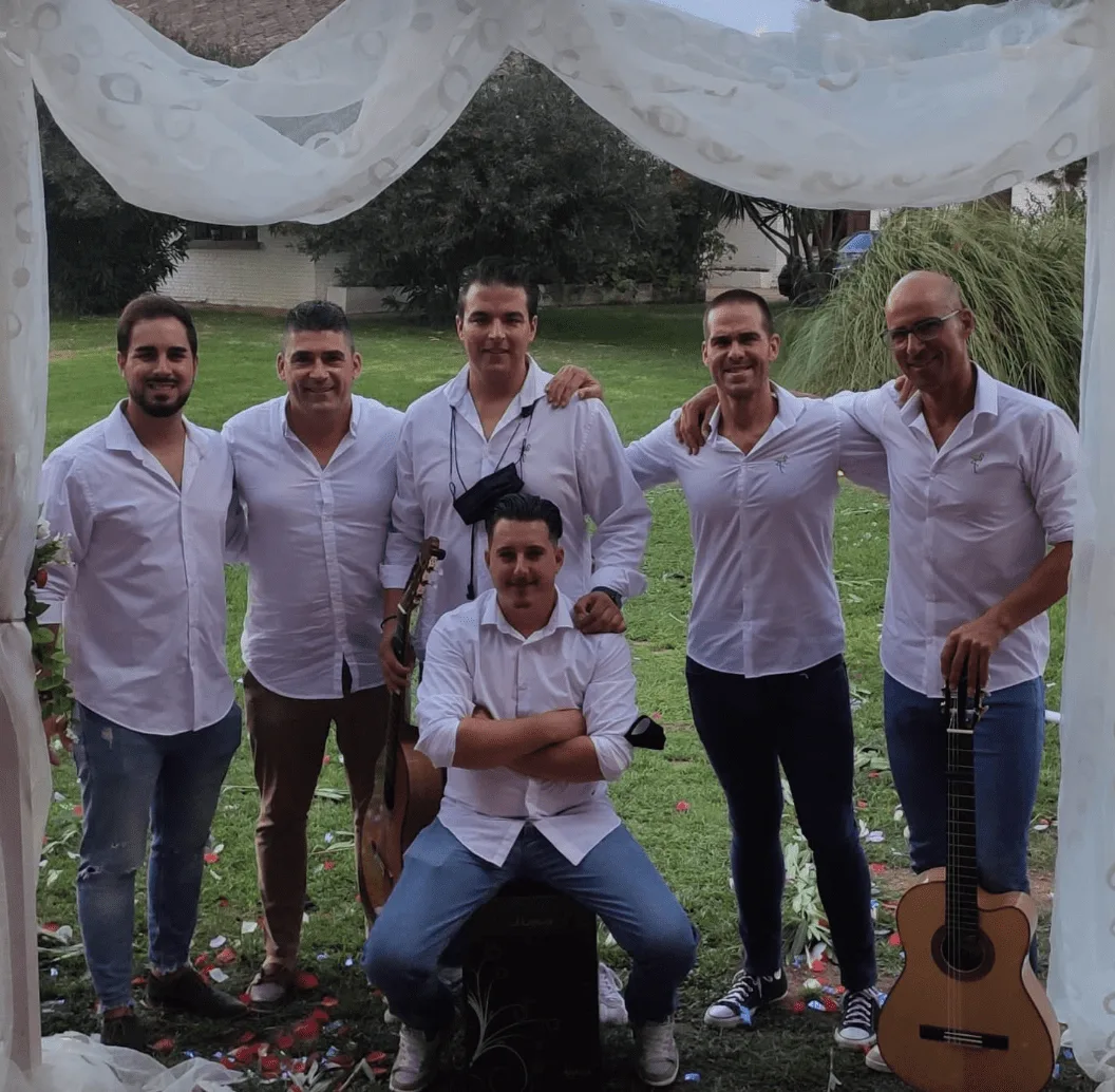 Cinco músicos posan juntos bajo un arco de tela blanca en un jardín, vestidos con camisas blancas; uno está sentado sobre un cajón y se ven dos guitarras apoyadas a los lados, una escena típica de rumba y flamenquito para bodas con buen rollo en Badajoz.