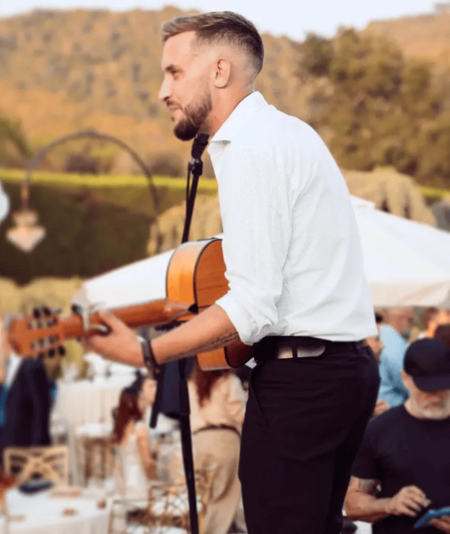 Músico de perfil con camisa blanca tocando una guitarra acústica junto a un micrófono, en una celebración al aire libre con invitados y mesas al fondo; imagen pensada para un dúo de flamenquito y rumba catalana para bodas en Barcelona.