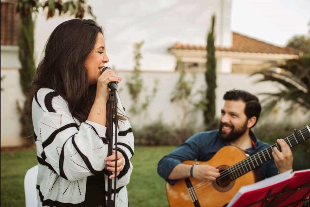 Cantora de cabelo escuro a segurar um microfone num jardim, enquanto um guitarrista sentado toca violao espanhol e sorri para ela; ao fundo ve se uma casa e arvores, num ambiente de celebracao ideal para flamenquito e rumba fusao para casamentos em Cordoba em formato trio.