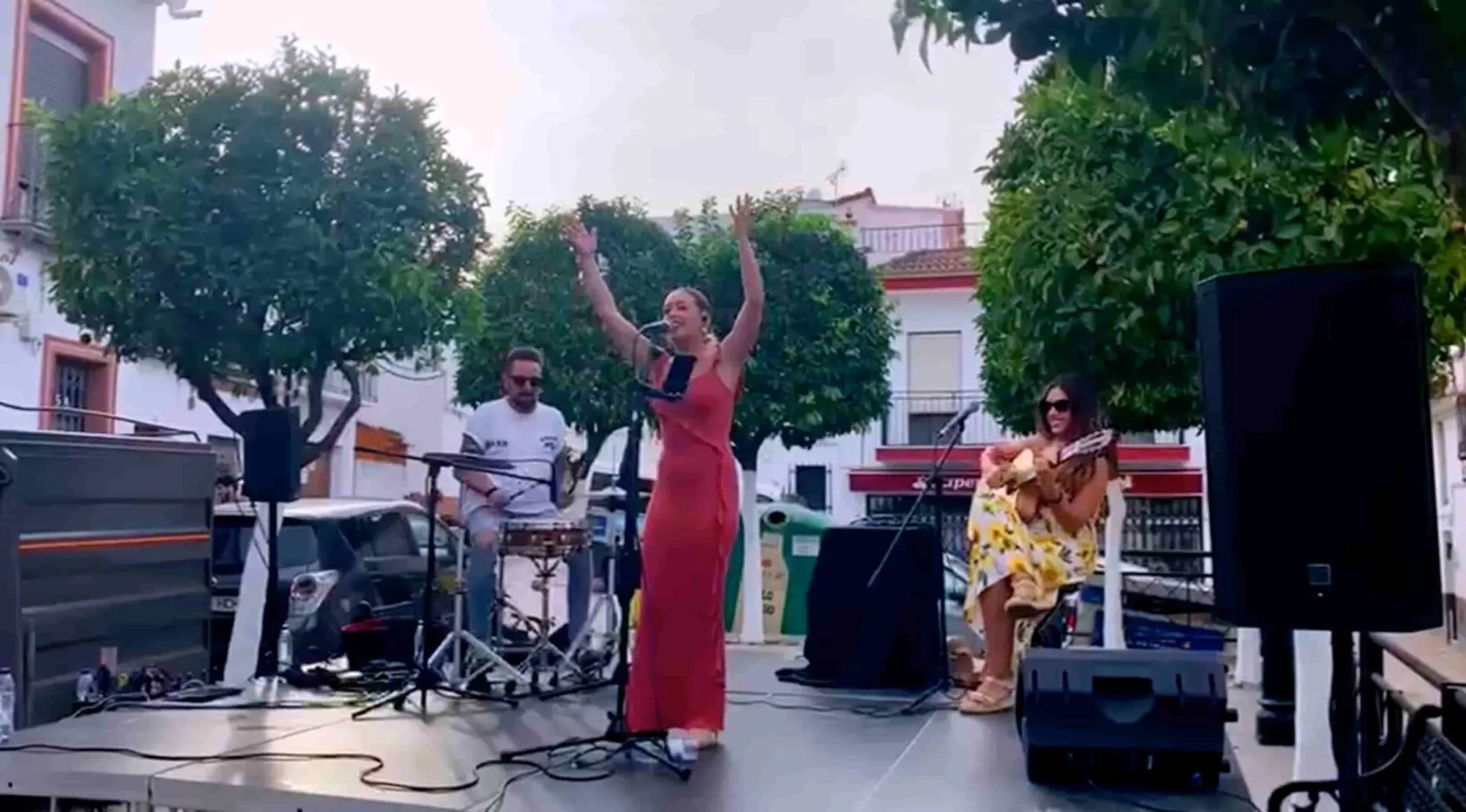 En un escenario al aire libre, una cantante con vestido rojo canta al micrófono con los brazos en alto; a su lado, una guitarrista sentada toca guitarra española y detrás un percusionista toca una caja. La escena muestra un trío de flamenquito y rumba, ideal para tu boda en Córdoba.