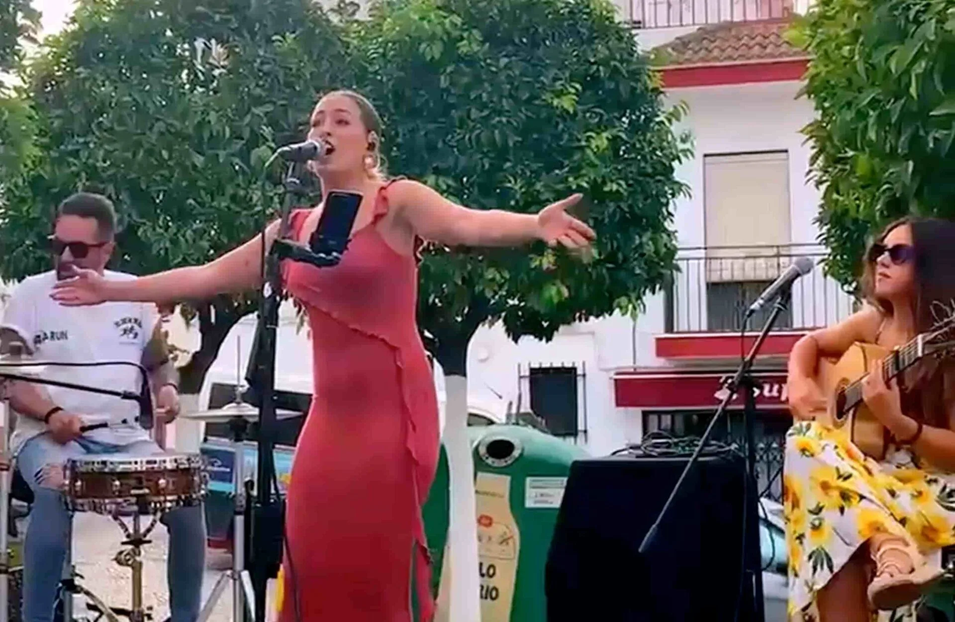 En un escenario al aire libre, una cantante con vestido rojo canta frente a un micrófono con los brazos abiertos, acompañada por un percusionista a la izquierda y una guitarrista sentada a la derecha; una imagen de un trío con flamenquito y rumba para tu boda en Córdoba.