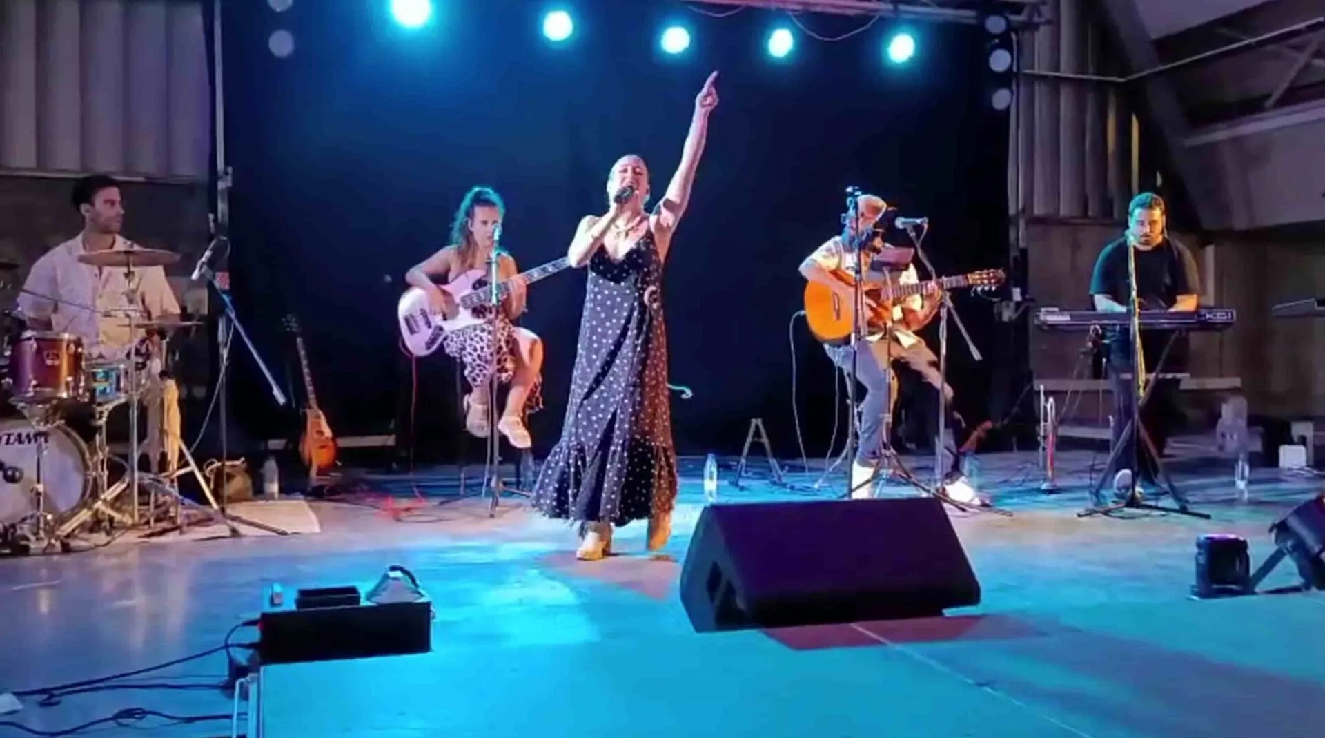 Escenario con iluminación azul: una cantante en el centro con micrófono y el brazo levantado, acompañada por batería a la izquierda, una bajista sentada, un guitarrista acústico y un teclista a la derecha; una escena ideal para un trío con flamenquito y rumba para tu boda en Córdoba.