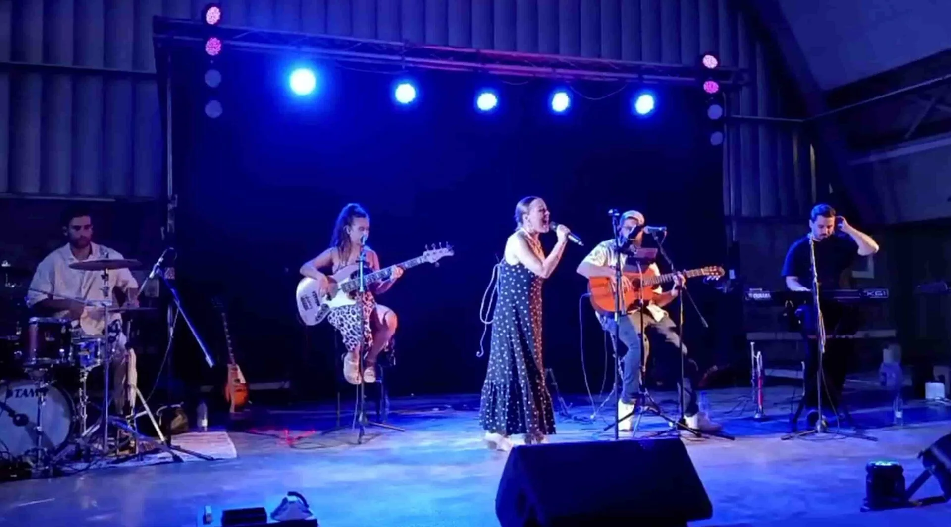 Grupo sobre un escenario con luces azules: una cantante en el centro con micrófono, una bajista sentada a su izquierda, un guitarrista sentado a la derecha y un batería al fondo; imagen apta para ilustrar un trío con flamenquito y rumba para tu boda en Córdoba.
