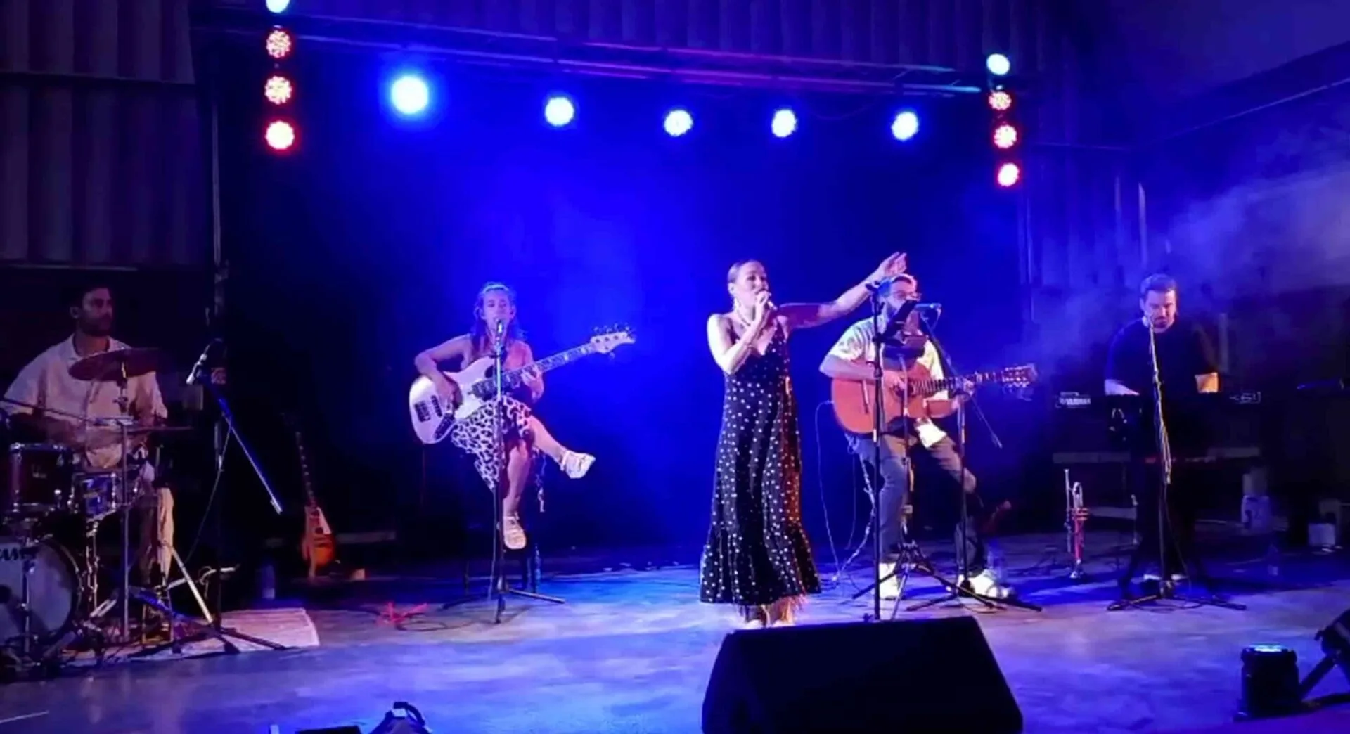 Grupo de música española sobre un escenario con luces azules: una cantante en el centro con micrófono y el brazo en alto, acompañada por batería, bajista, guitarrista acústico y teclista; una imagen pensada para quien busca un trío con flamenquito y rumba para tu boda en Córdoba.