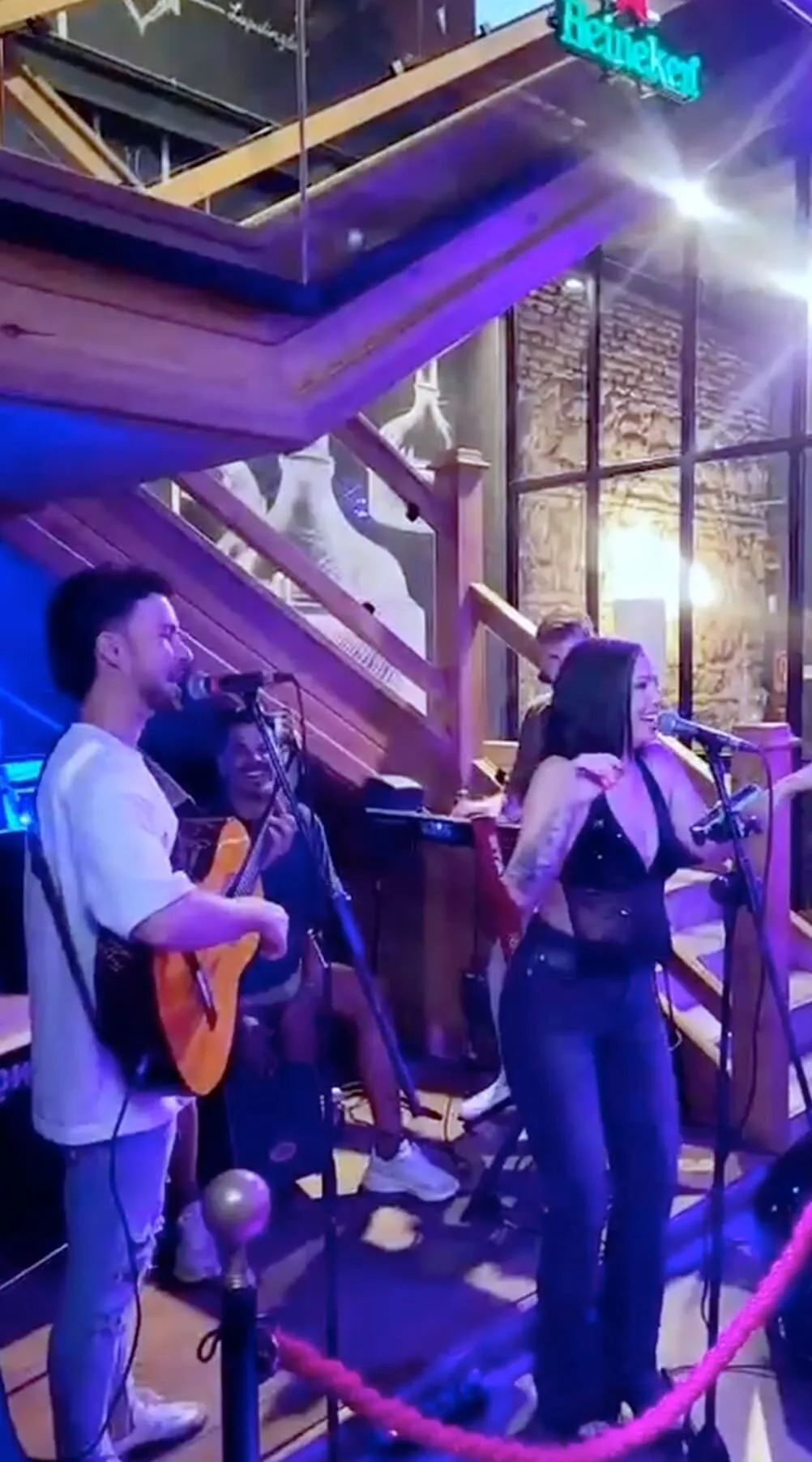 Pequeño escenario interior con un trío tocando: un guitarrista cantando al micrófono, una cantante frente a otro micro con gesto animado y un tercer músico al fondo; iluminación morada y escaleras de madera detrás. Imagen ideal para ilustrar un trío pop flamenco en Córdoba para darle vida al tardeo de boda.