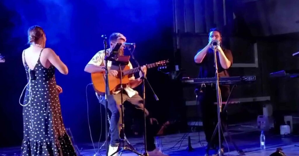 Num palco com luz azul, uma mulher de costas com vestido preto às bolinhas observa um músico sentado a tocar guitarra com microfone à frente, enquanto outro músico toca trompete ao lado de um teclado. Imagem de um trio com flamenquito e rumba para a sua boda em Córdoba.