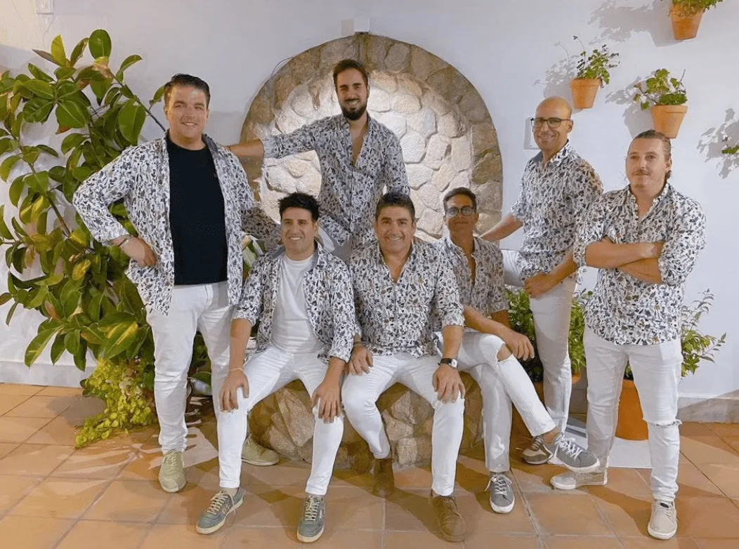Foto de un grupo de seis hombres posando juntos en una terraza con pared blanca y arco de piedra, rodeados de plantas y macetas; llevan camisas estampadas y pantalones blancos, una imagen típica para promocionar rumba y flamenquito para bodas con buen rollo en Badajoz.