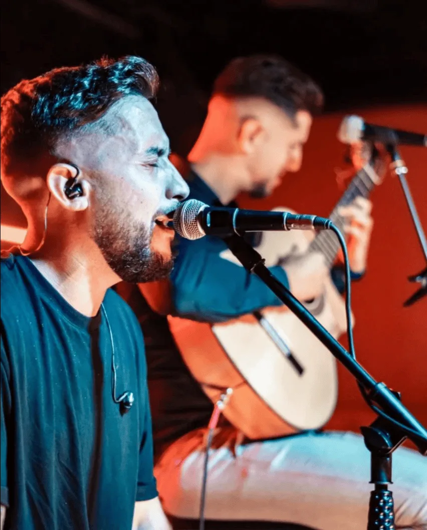 Primer plano de un cantante con barba y auricular cantando en un micrófono, mientras al fondo, desenfocado, otro músico toca una guitarra española; imagen de un dúo de flamenquito y rumba catalana para bodas en Barcelona.