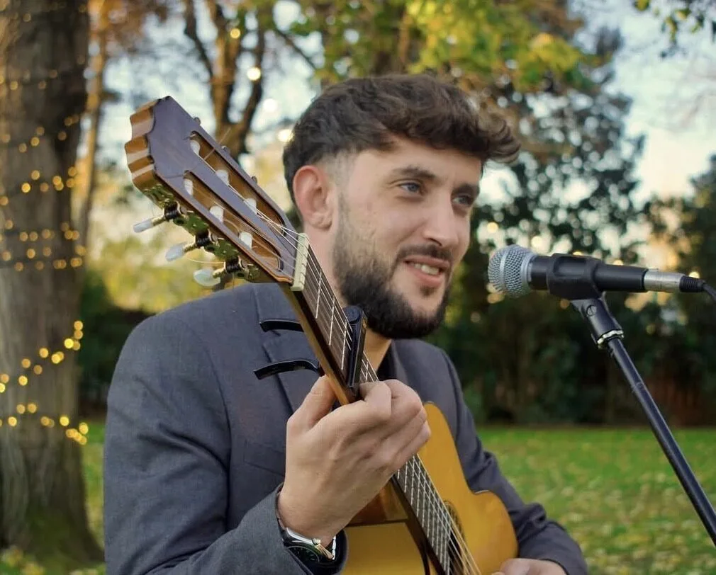 Primer plano de un músico con guitarra española junto a un micrófono, con árboles y luces cálidas desenfocadas al fondo, en un ambiente exterior tipo celebración; imagen representativa de un dúo de flamenquito y rumba catalana para bodas en Barcelona.