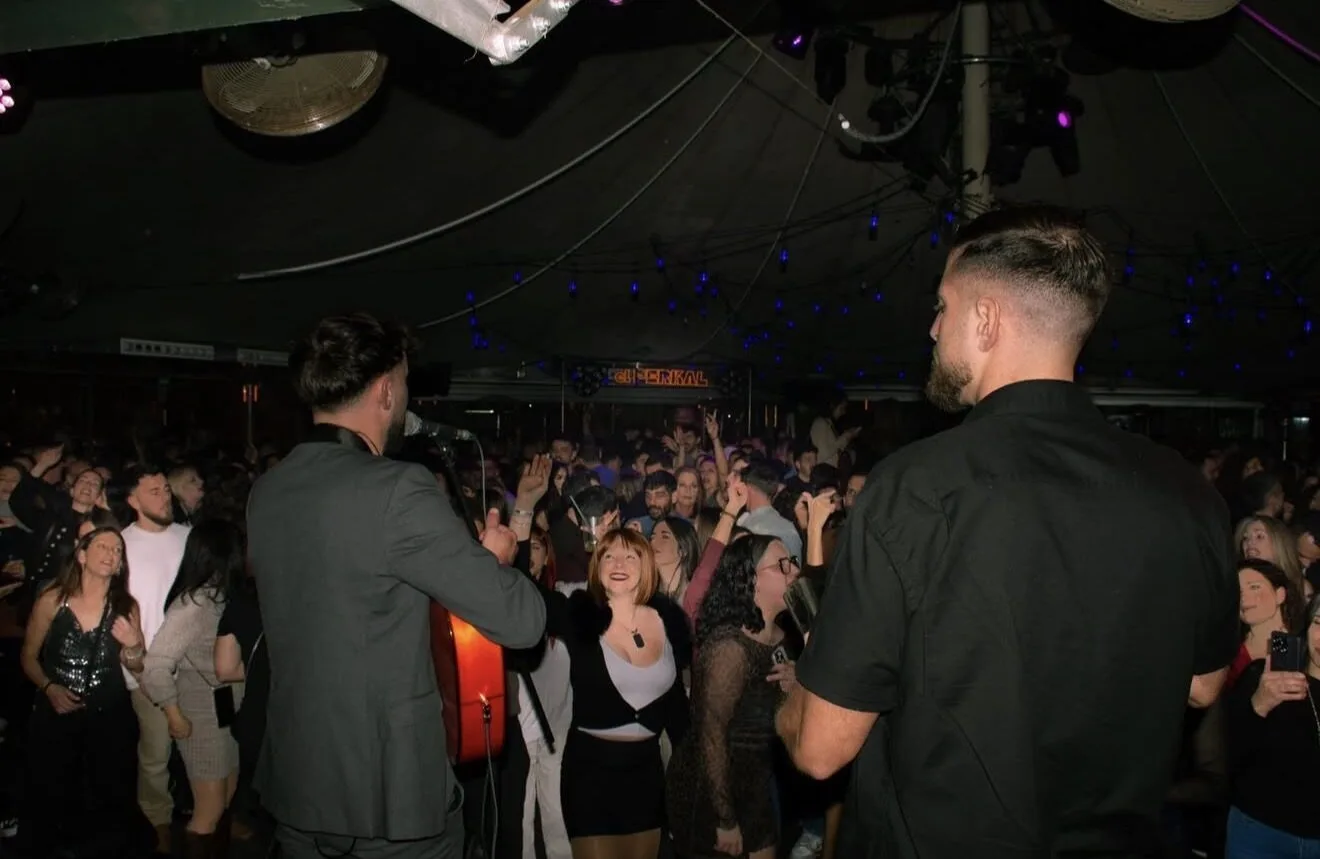 Vista desde el escenario: dos músicos de espaldas, uno con guitarra y micrófono y otro junto a él, frente a una sala llena de gente con las manos levantadas y luces tenues, en un ambiente de celebración. Imagen asociada a un dúo de flamenquito y rumba catalana para bodas en Barcelona.