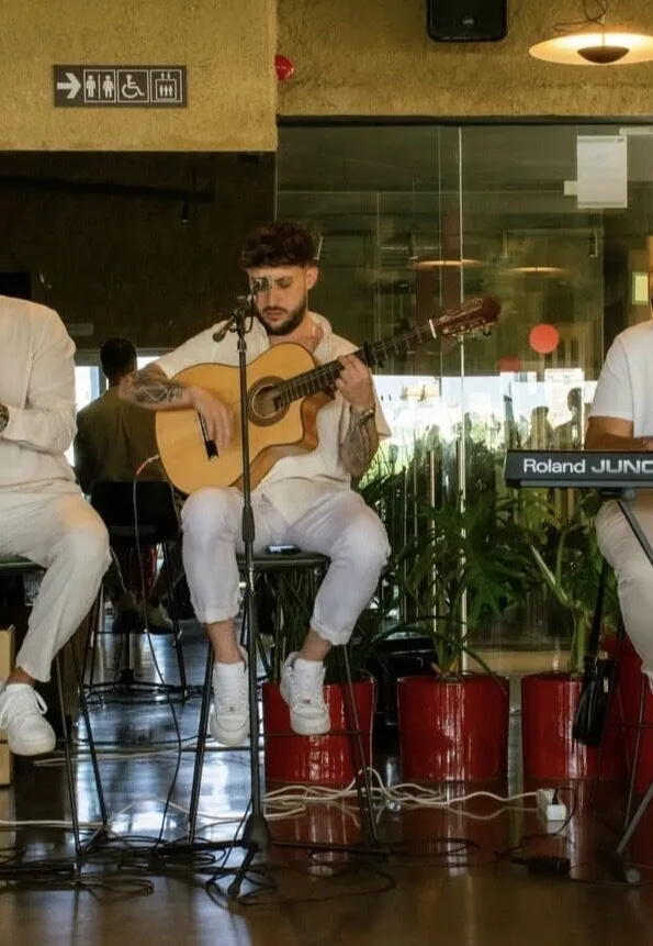 Músico sentado tocando una guitarra española frente a un micrófono, con cables en el suelo; a los lados se ven parcialmente otros intérpretes, incluido un teclado, en un espacio interior con cristaleras. Imagen de un dúo de flamenquito y rumba catalana para bodas en Barcelona.