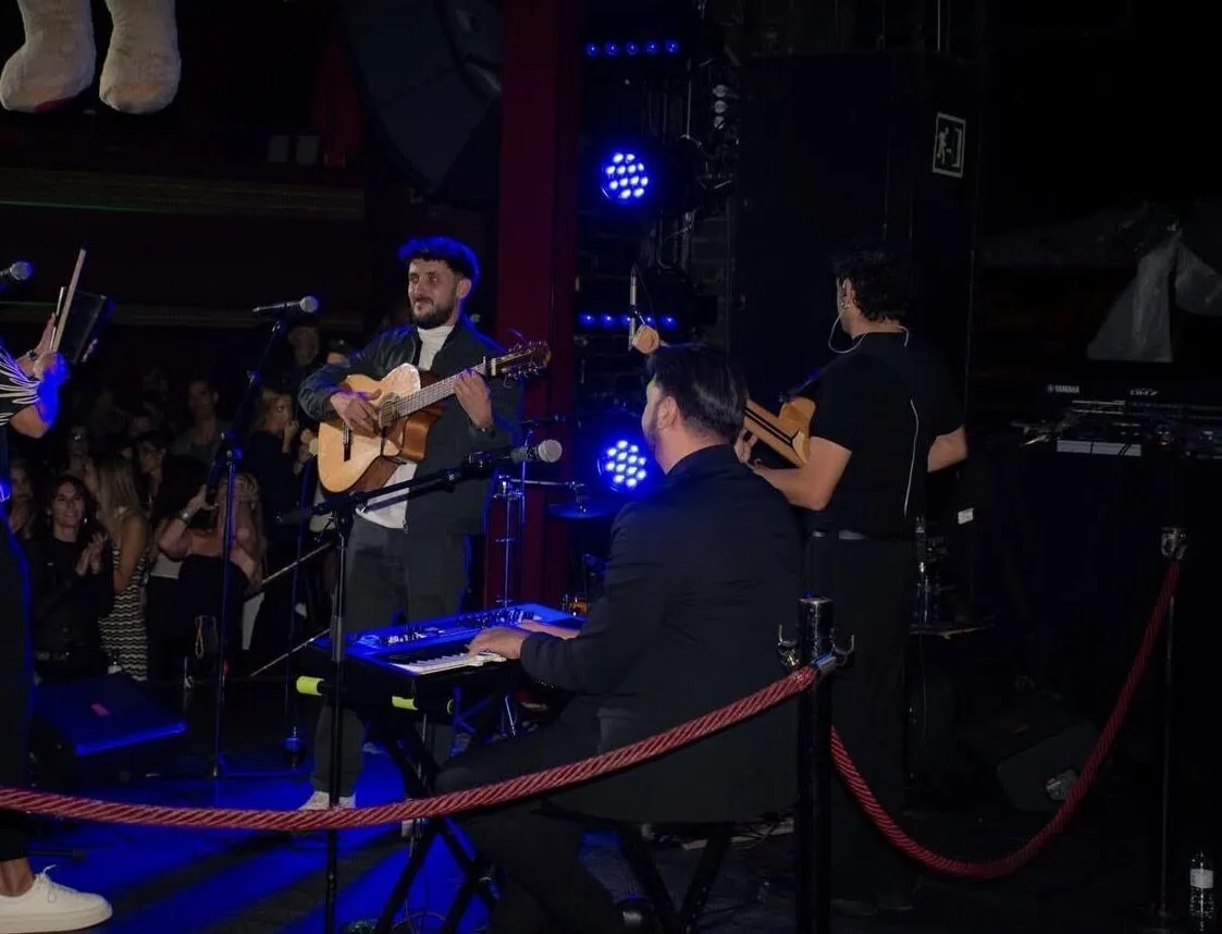 En un pequeño escenario interior, un guitarrista de pie toca una guitarra española frente a un público muy cercano; a su lado se ve otro músico con guitarra de espaldas y, en primer plano, un pianista sentado. La escena está iluminada con focos azules y sugiere un dúo de flamenquito y rumba catalana para bodas en Barcelona.