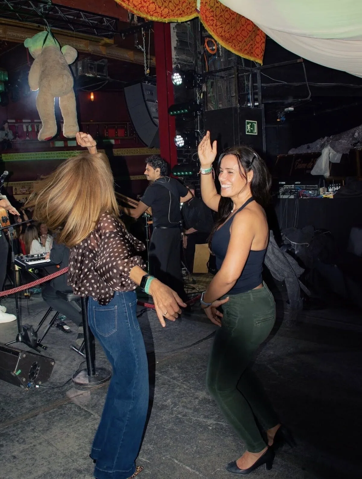 Dos mujeres bailan sonrientes con los brazos levantados cerca de un pequeño escenario con músicos y focos, en un local interior; ambiente de fiesta con música española, ideal para un dúo de flamenquito y rumba catalana para bodas en Barcelona.