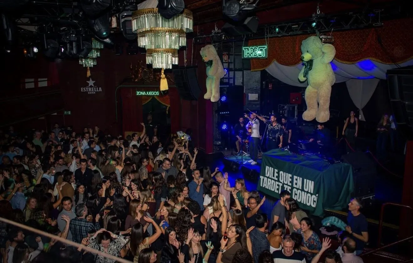 Sala de fiesta llena de gente bailando delante de un escenario iluminado; arriba cuelgan dos osos de peluche grandes y se ven carteles de neón. En el escenario hay varios músicos, una imagen que encaja con un dúo de flamenquito y rumba catalana para bodas en Barcelona.