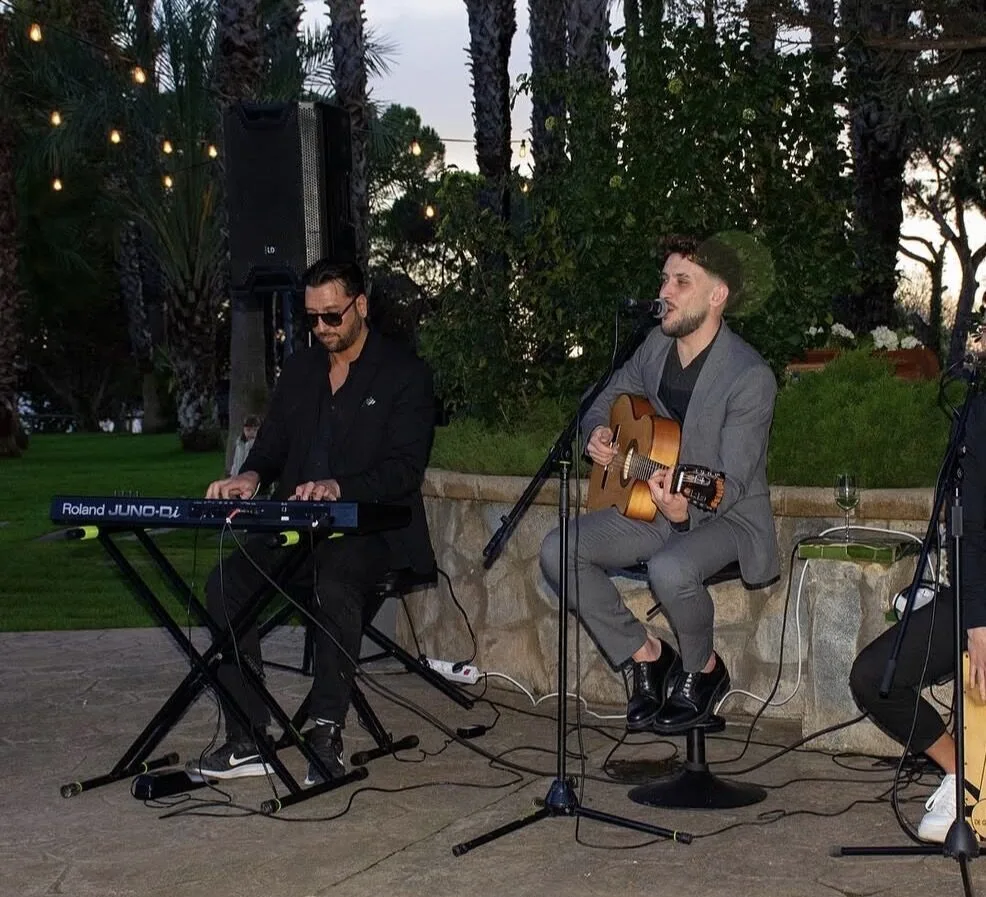 Dos músicos actuando al aire libre en un jardín al atardecer: uno toca un teclado y el otro canta a un micrófono mientras toca guitarra española sentado en un taburete; hay un altavoz, cables y luces colgantes entre los árboles, una escena típica de un dúo de flamenquito y rumba catalana para bodas en Barcelona.