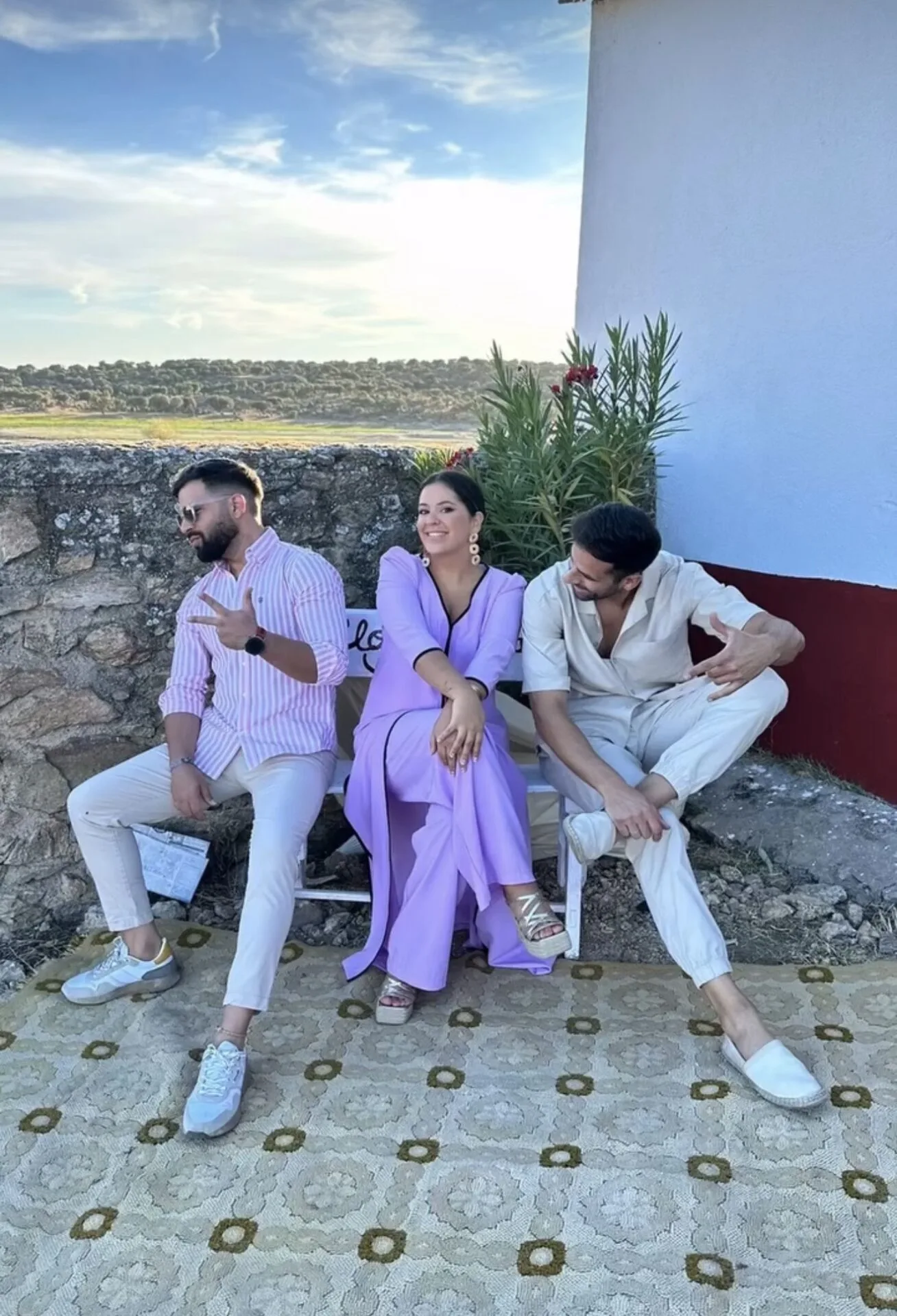 Tres personas sentadas en un banco exterior junto a una pared blanca: una mujer con vestido lila en el centro sonriendo a cámara y dos hombres a los lados con ropa clara, uno con gafas de sol. Al fondo se ve un muro de piedra, plantas altas y un paisaje abierto bajo un cielo azul, en un ambiente que encaja con flamenquito y rumba fusión para bodas en Córdoba en formato trío.
