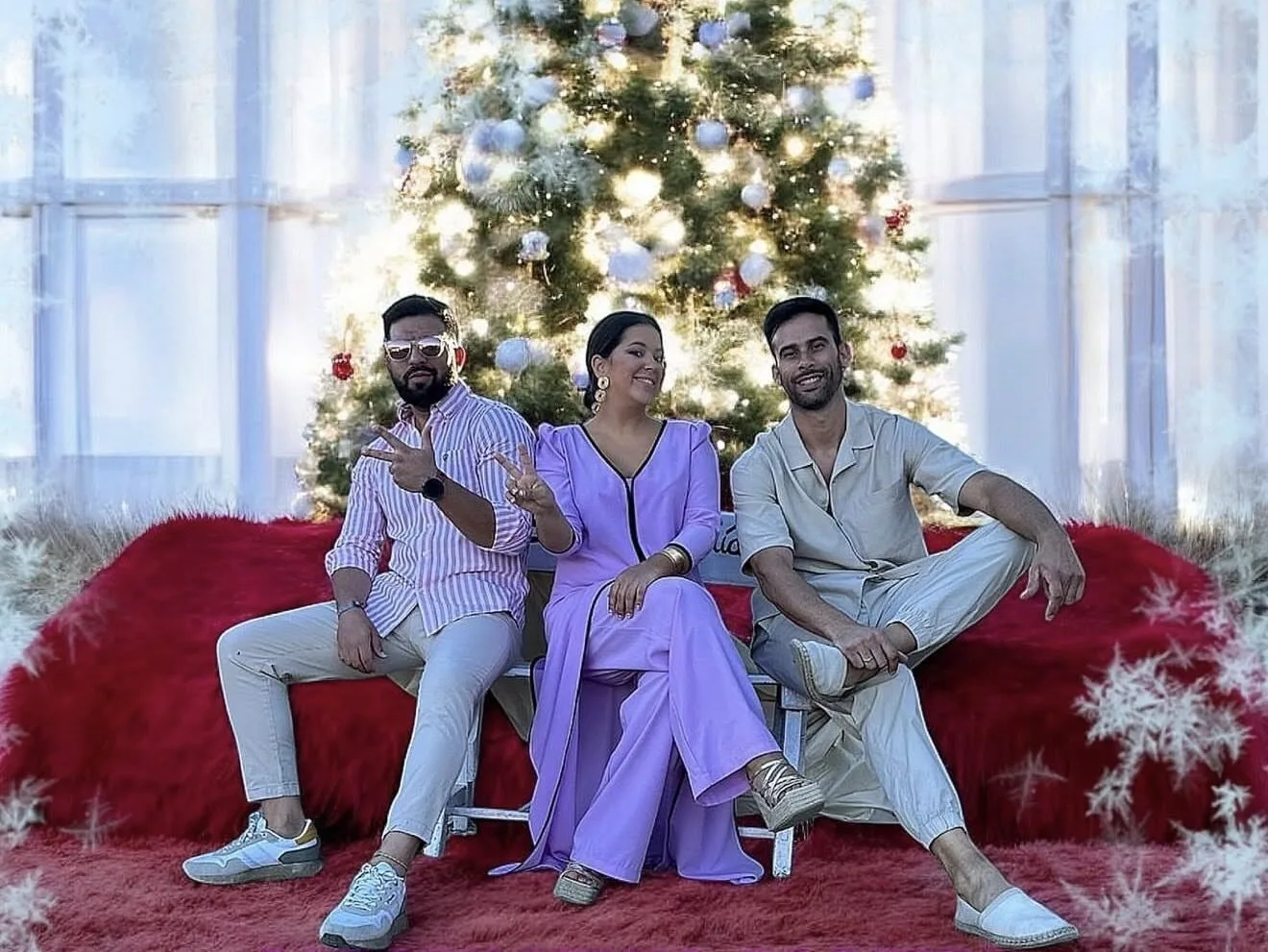 Tres músicos posan sentados en un banco delante de un gran árbol de Navidad iluminado, sobre una alfombra roja. La persona del centro viste de lila y los dos acompañantes llevan ropa clara; uno hace el gesto de la paz. Imagen de un trío de flamenquito y rumba fusión para bodas en Córdoba.