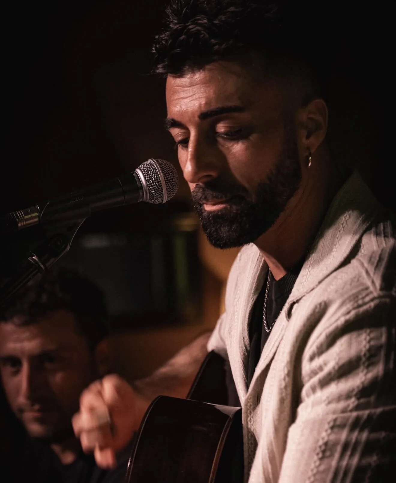 Primer plano de un cantante con barba y pendiente, concentrado frente a un micrófono mientras toca una guitarra; al fondo aparece otra persona desenfocada. La escena sugiere un ambiente íntimo y cercano, ideal para rumba catalana y mestizaje para bodas con buen rollo en Barcelona.