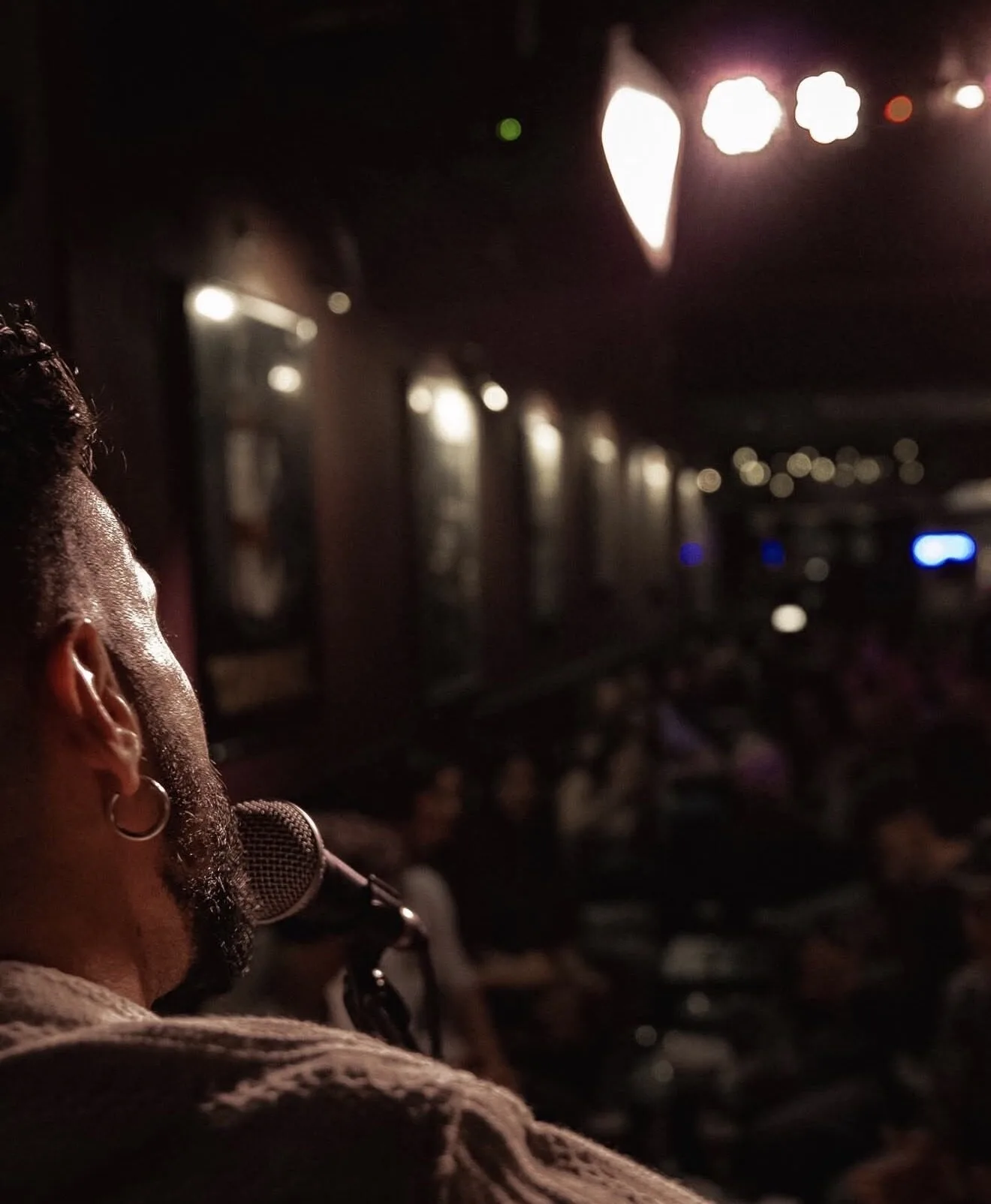 Plano corto desde detrás de un cantante con pendiente en la oreja, junto a un micrófono, mirando hacia una sala con público desenfocado y luces cálidas; imagen que sugiere rumba catalana y mestizaje para bodas con buen rollo en Barcelona.