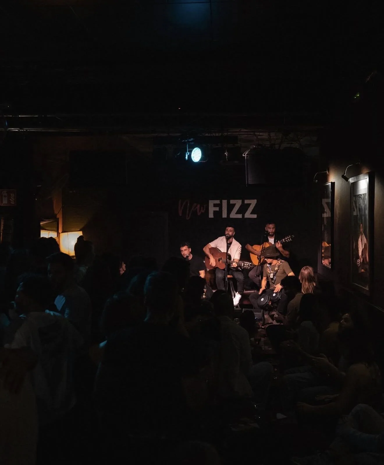 Escenario pequeño en una sala oscura con un grupo de rumba tocando: dos músicos con guitarra sentados y un percusionista con cajón, mientras el público llena la parte inferior de la imagen. Sobre el fondo se ve el rótulo “FIZZ” y una luz azul ilumina la escena, con un ambiente de rumba catalana y mestizaje ideal para bodas con buen rollo en Barcelona.