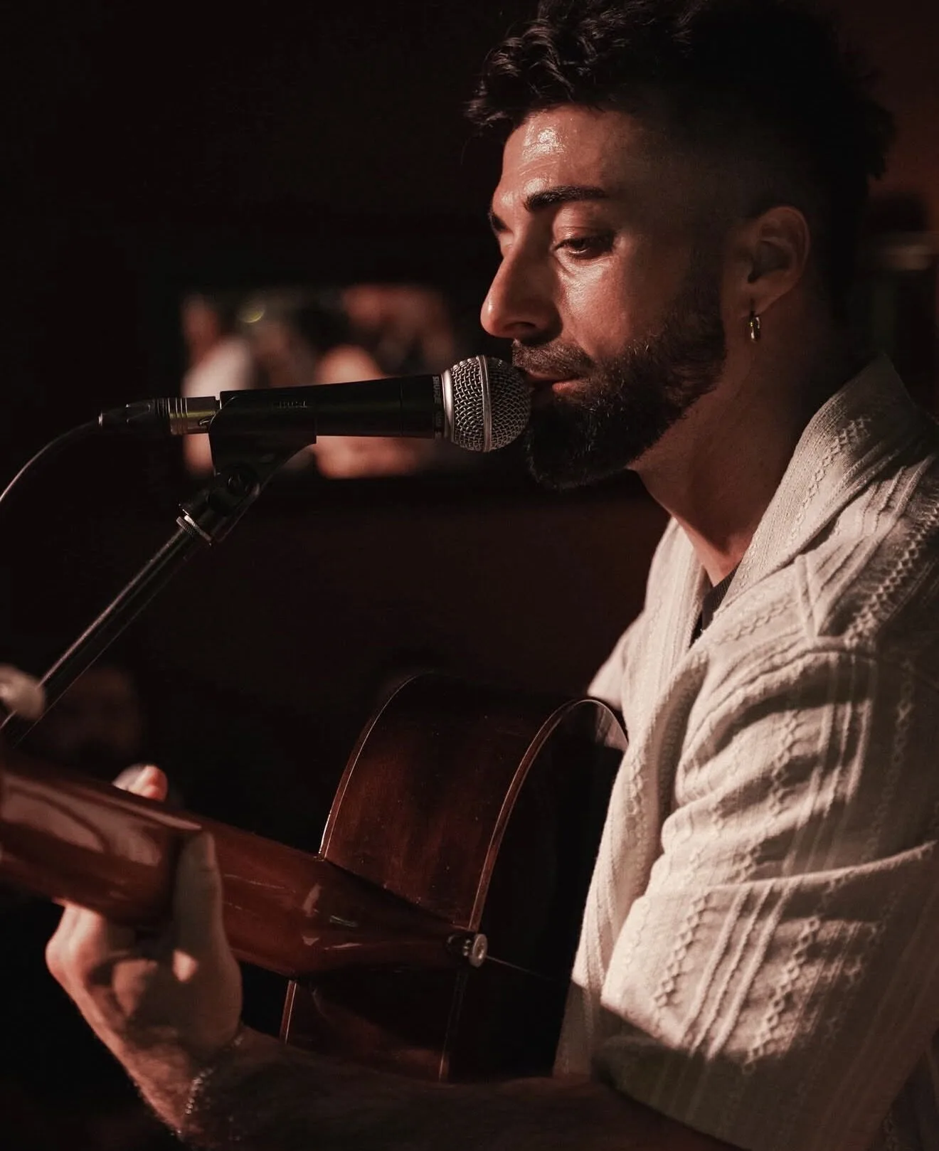 Primer plano de un músico cantando en un micrófono mientras toca una guitarra española; lleva barba y un pendiente, con luz cálida y el público desenfocado al fondo, una escena íntima de rumba catalana y mestizaje para bodas con buen rollo en Barcelona.