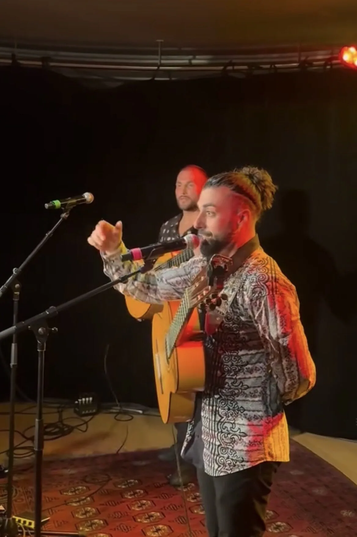 Dos músicos sobre un pequeño escenario con fondo negro: en primer plano, un cantante con barba y coleta habla al micrófono mientras lleva una guitarra española colgada; detrás, otro músico sostiene una guitarra. La iluminación es cálida con tonos rojos, ideal para un ambiente de rumba catalana y mestizaje para bodas con buen rollo en Barcelona.