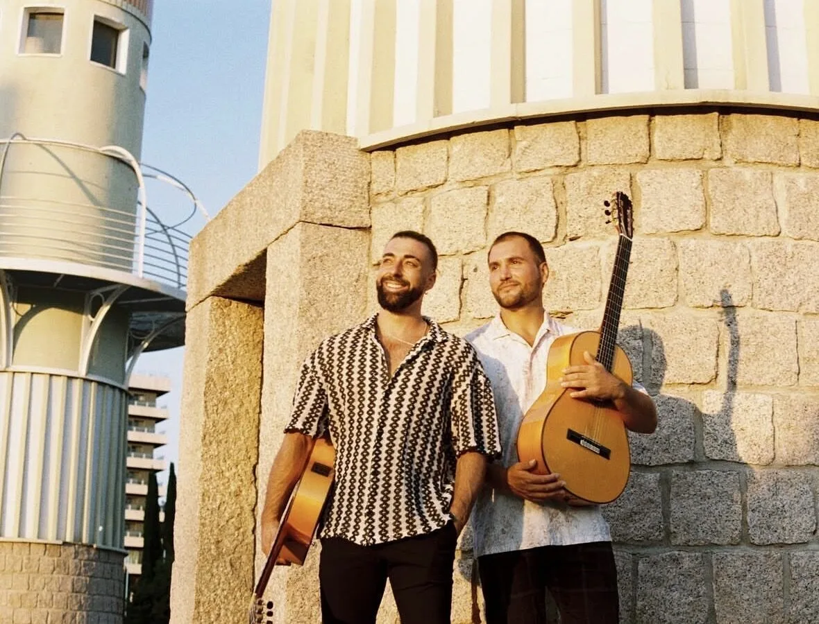 Dos músicos posan de pie junto a una pared de piedra, con dos guitarras españolas en las manos. Están al aire libre con luz cálida, mirando hacia un lado y sonriendo; la escena transmite un estilo cercano de rumba catalana y mestizaje para bodas con buen rollo en Barcelona.