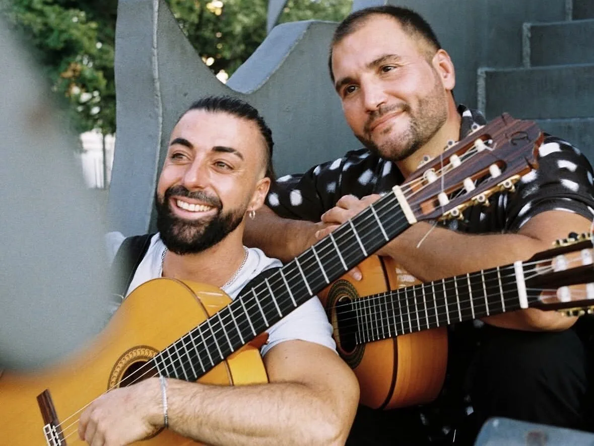 Dos músicos sonrientes posan sentados con guitarras españolas, uno en primer plano y otro detrás apoyado en su instrumento; una imagen cálida que encaja con rumba catalana y mestizaje para bodas con buen rollo en Barcelona.