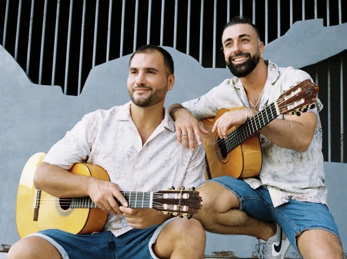 Dos músicos sentados al aire libre con guitarras españolas en el regazo; uno mira hacia un lado y el otro sonríe, con su brazo apoyado en el hombro del compañero, en un ambiente relajado que encaja con rumba catalana y mestizaje para bodas con buen rollo en Barcelona.