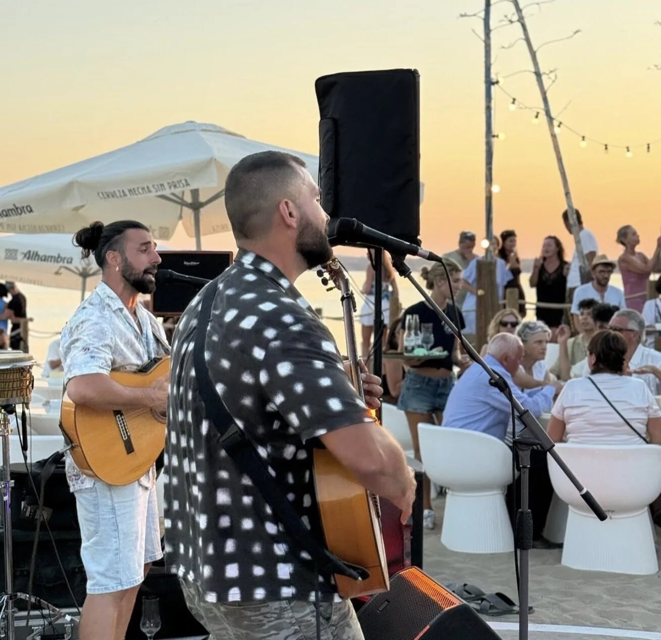 Dos músicos tocando guitarras y cantando con micrófonos en un chiringuito de playa al atardecer, con público sentado y de pie detrás; ambiente relajado de rumba catalana y mestizaje para bodas con buen rollo en Barcelona.