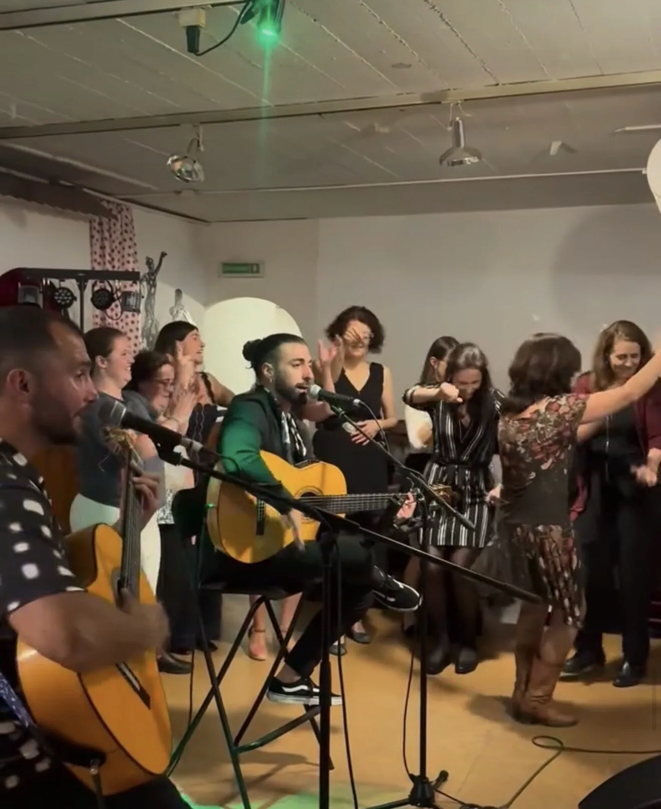 En una sala interior, dos músicos con guitarras españolas tocan sentados junto a micrófonos mientras varias personas alrededor aplauden y bailan muy cerca; una mujer en primer plano se mueve con los brazos en alto, creando un ambiente de rumba catalana y mestizaje ideal para bodas con buen rollo en Barcelona.