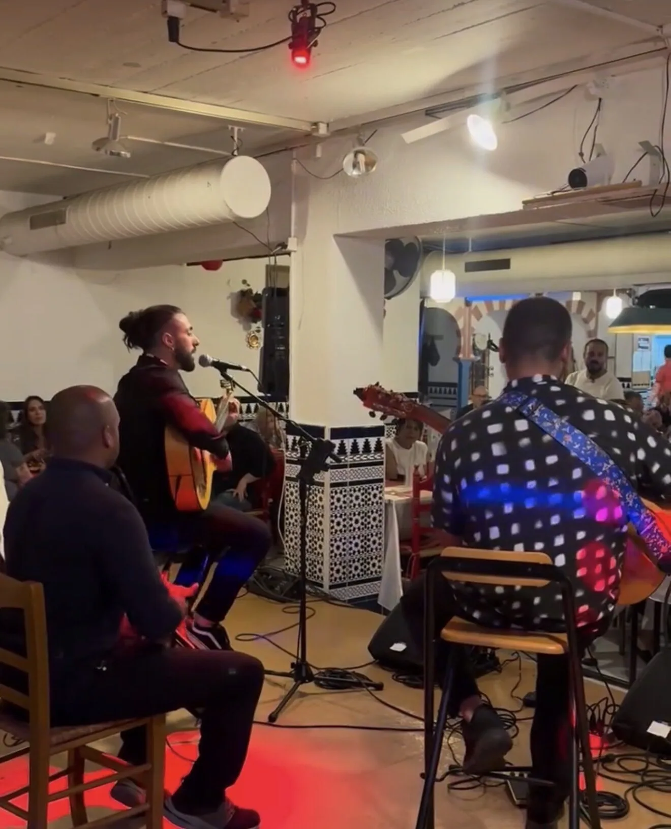 Dos músicos tocando guitarras en un local con mesas: uno canta al micrófono mientras el público escucha sentado alrededor, una escena cercana ideal para rumba catalana y mestizaje para bodas con buen rollo en Barcelona.