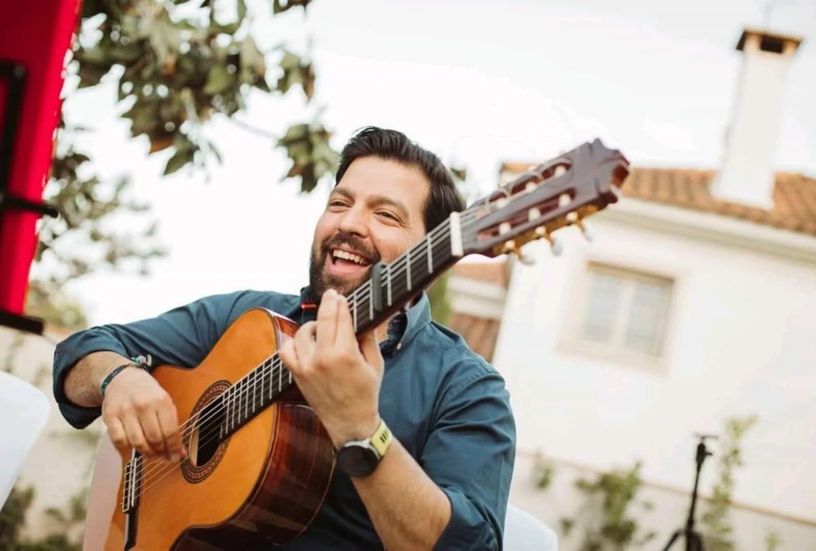 Primer plano de un guitarrista sonriendo mientras toca una guitarra española, en un entorno exterior con una fachada blanca al fondo; imagen ideal para ilustrar flamenquito y rumba fusión para bodas en Córdoba, en formato trío.