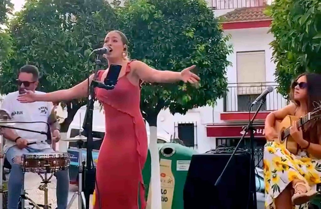 Cantora de vestido vermelho canta ao microfone com os bracos abertos, acompanhada por um percussionista e uma guitarrista sentada com violao, num palco ao ar livre com arvores e uma fachada branca ao fundo, sugerindo um trio de flamenquito e rumba para tua boda em Cordoba